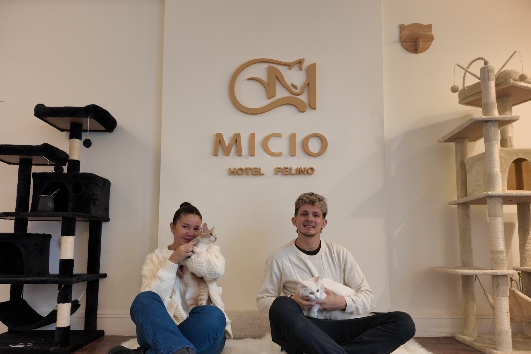 Paula y Juan sentados en la Sala Micio con dos gatos en brazos, frente al cartel de Micio Hotel Felino